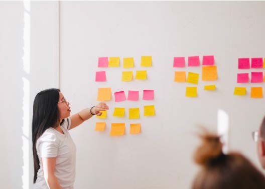 A woman with long dark hair points to a white board covered in yellow and pink post-it notes; cut off image of another person watching